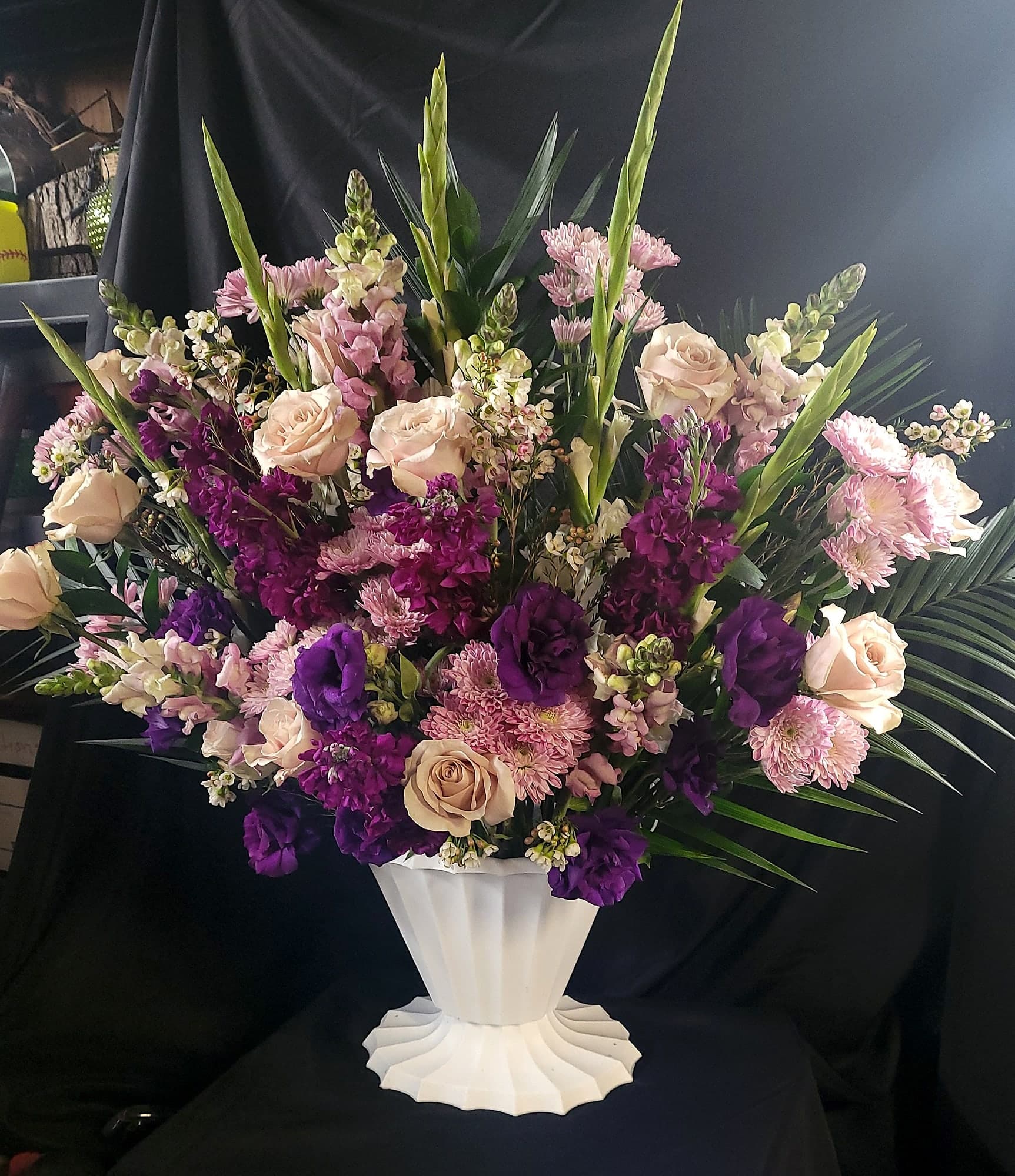 Tall formal floral arrangement in a white pedestal urn, filled with purple, pink, and cream roses, stock, chrysanthemums, and gladiolus against a dark backdrop.