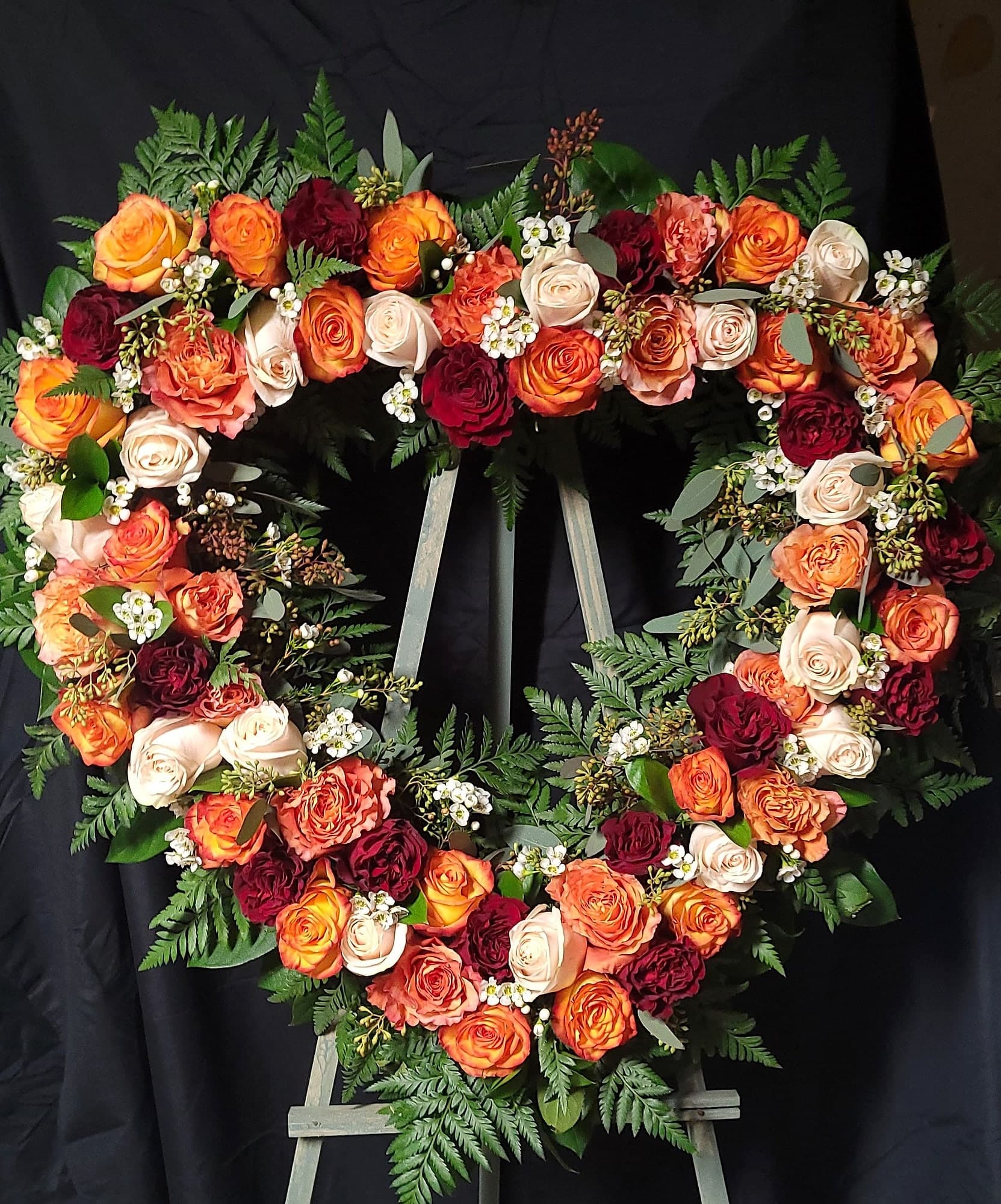 Heart-shaped tribute wreath of orange, cream, and burgundy roses with white waxflower and green fern, on a wooden easel in front of a black cloth.