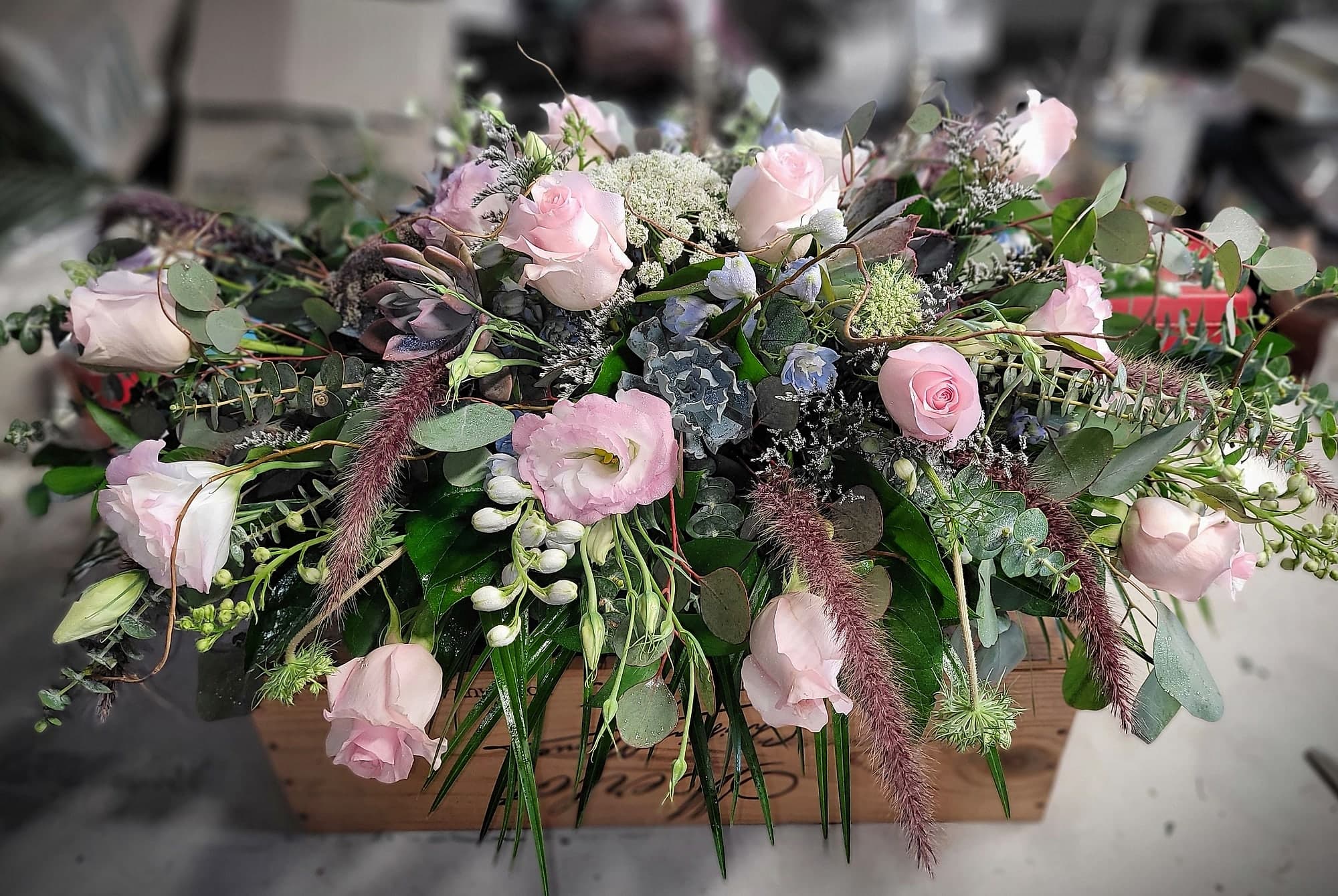 A long wooden box filled with an abundant low arrangement of pale pink roses, pink lisianthus, blue delphinium, eucalyptus, and dusty-rose grasses.