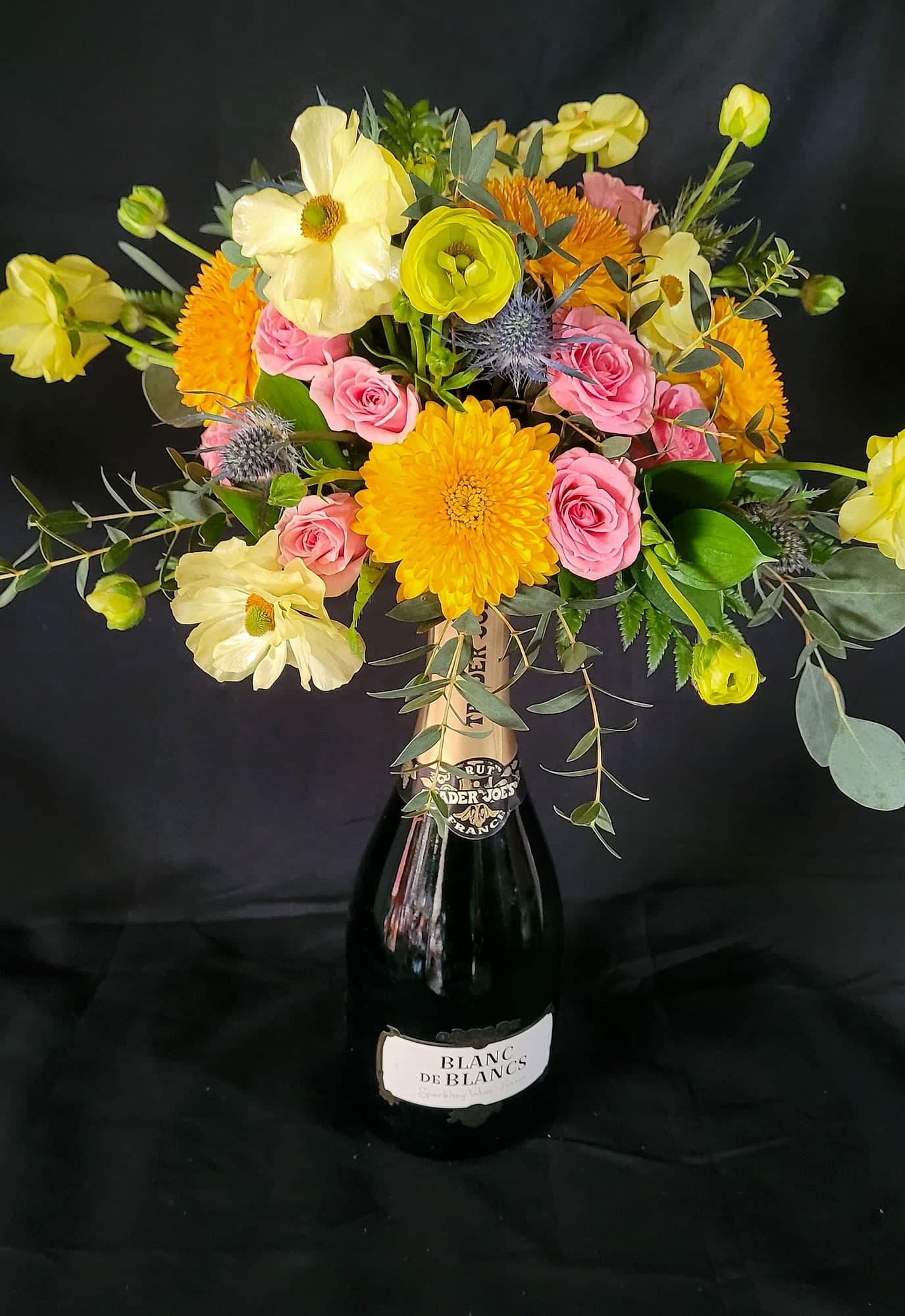 A colorful hand-tied bouquet with yellow poppies, orange chrysanthemums, pink spray roses, and blue thistle against a black backdrop.