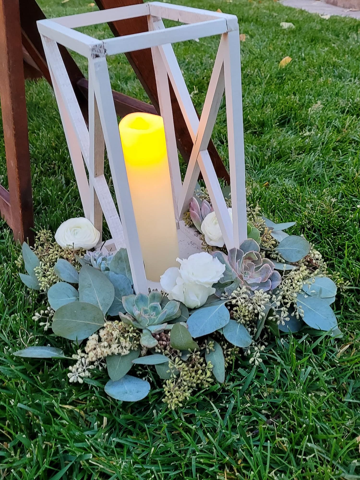 White wooden lantern holding a lit candle, surrounded by a wreath of green eucalyptus, gray succulents, and white flowers, resting on green grass outdoors.