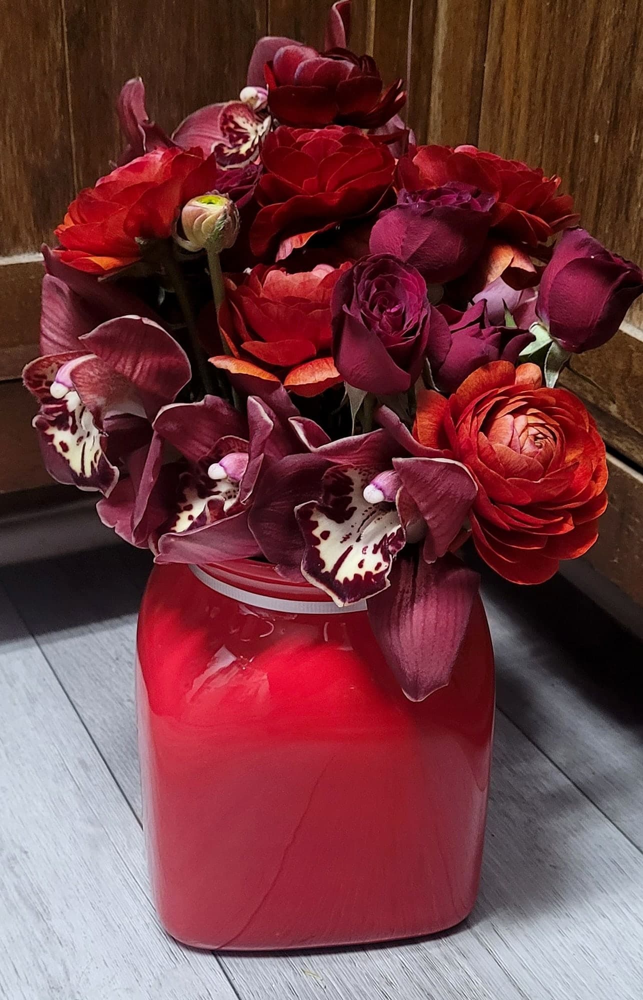 A red square vase holding a dense arrangement of deep red ranunculus, roses, and burgundy speckled orchids against dark wood paneling.