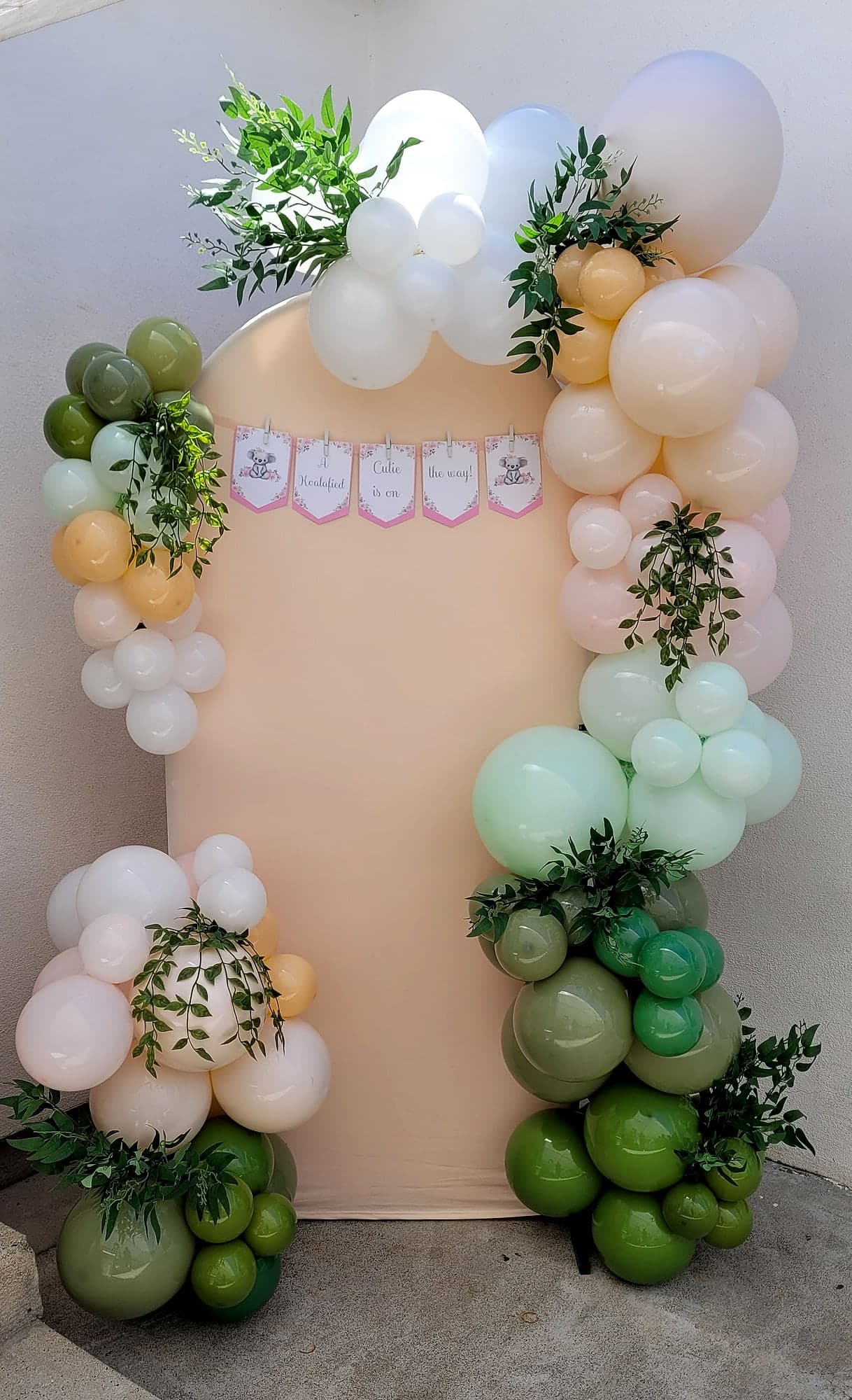 Arched tan backdrop framed by an organic balloon garland in sage green, peach, cream, and white with trailing greenery, and a banner reading Cutie is on the way.