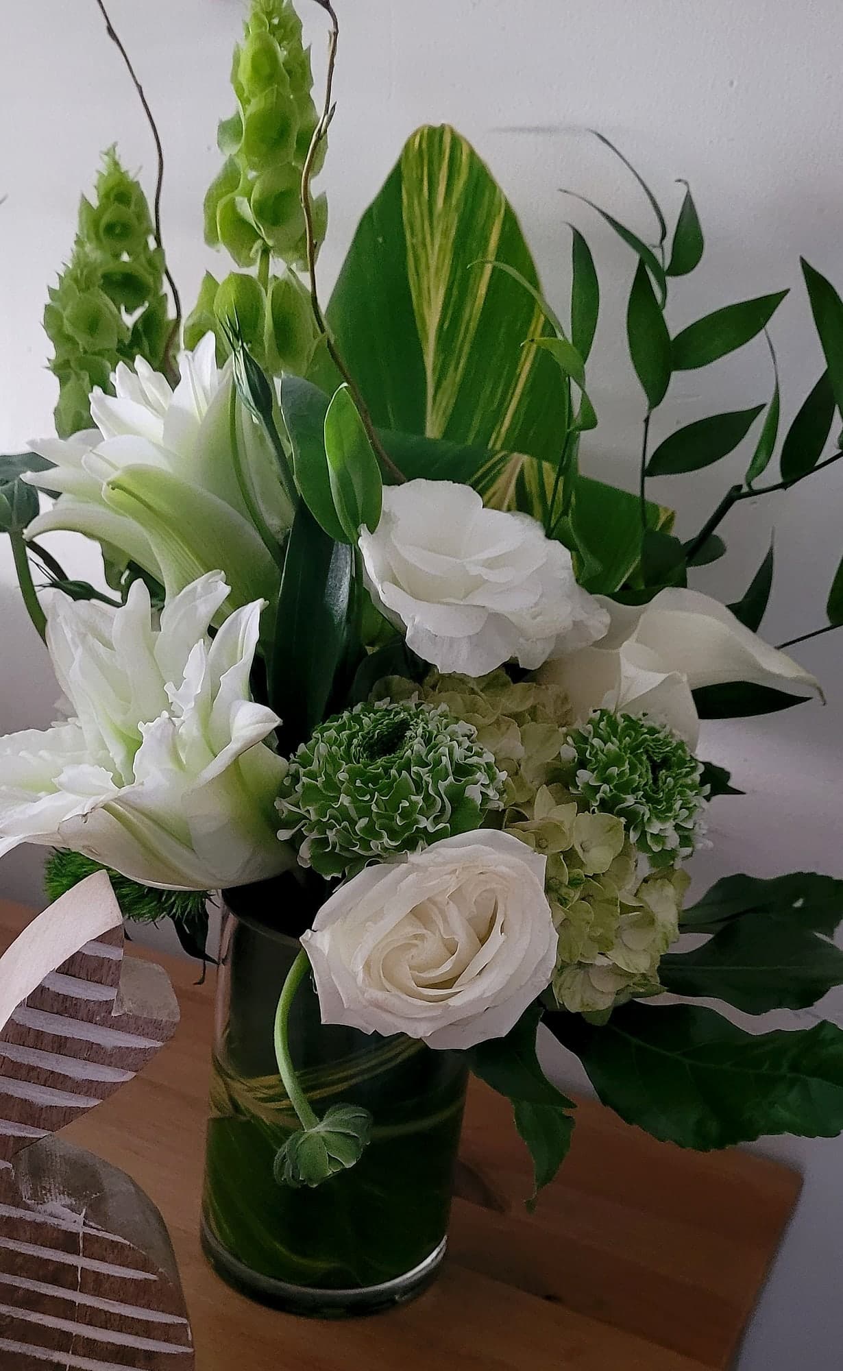 White roses, lisianthus, tulips, and green hydrangea with bells of Ireland arranged in a clear glass cylinder vase on a wooden table.