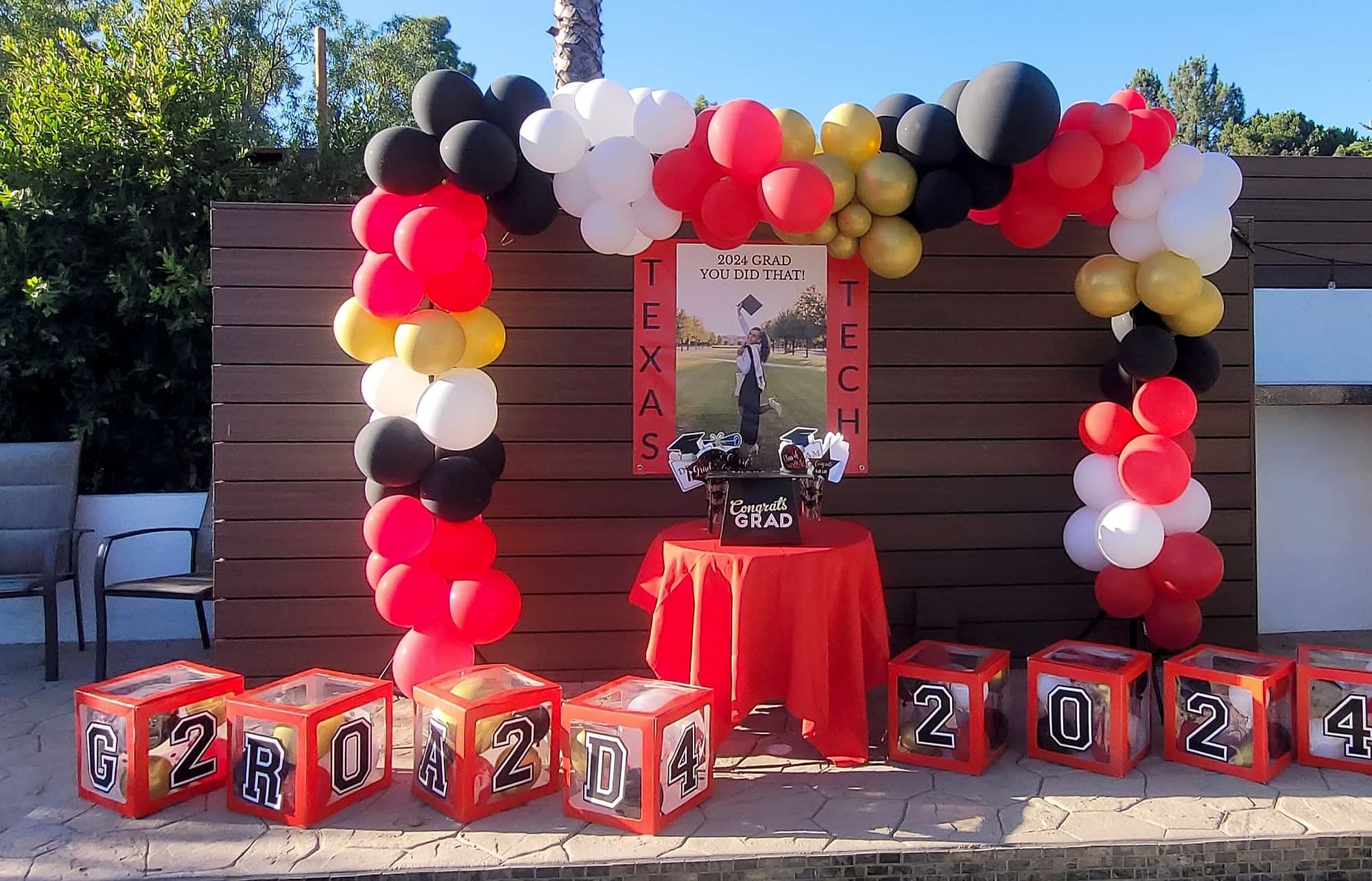 A large balloon garland arch in red, black, white, and gold over a wood fence, with a graduation photo banner, a red-draped table, and cube boxes spelling GRAD 2024.