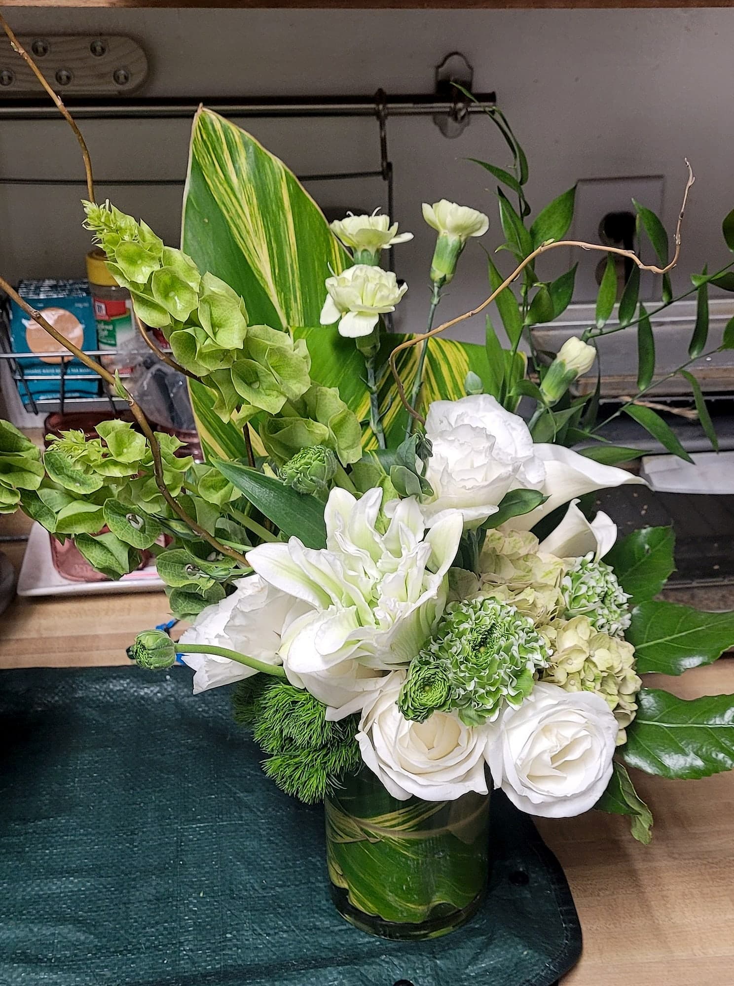 A floral arrangement of white roses, a white dahlia, green bells of Ireland, and tropical leaves in a clear glass vase.