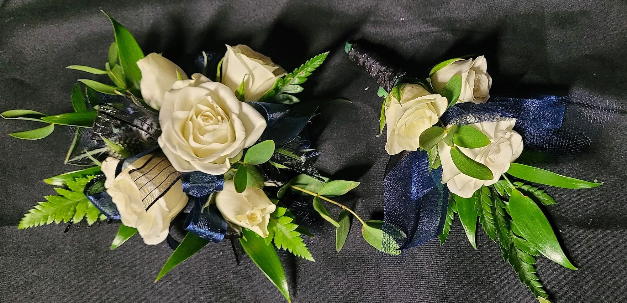 A wristlet corsage and matching boutonniere made of cream roses, navy blue ribbon, and green foliage arranged on a black satin backdrop.