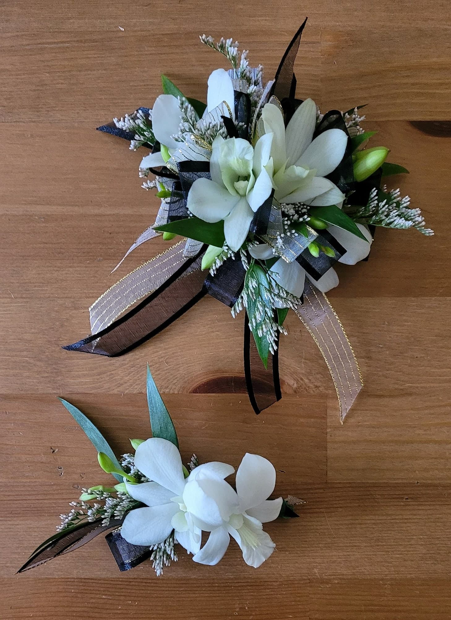 A wrist corsage and a boutonniere made of white orchids with greenery and black and gold ribbon, arranged on a wood table.