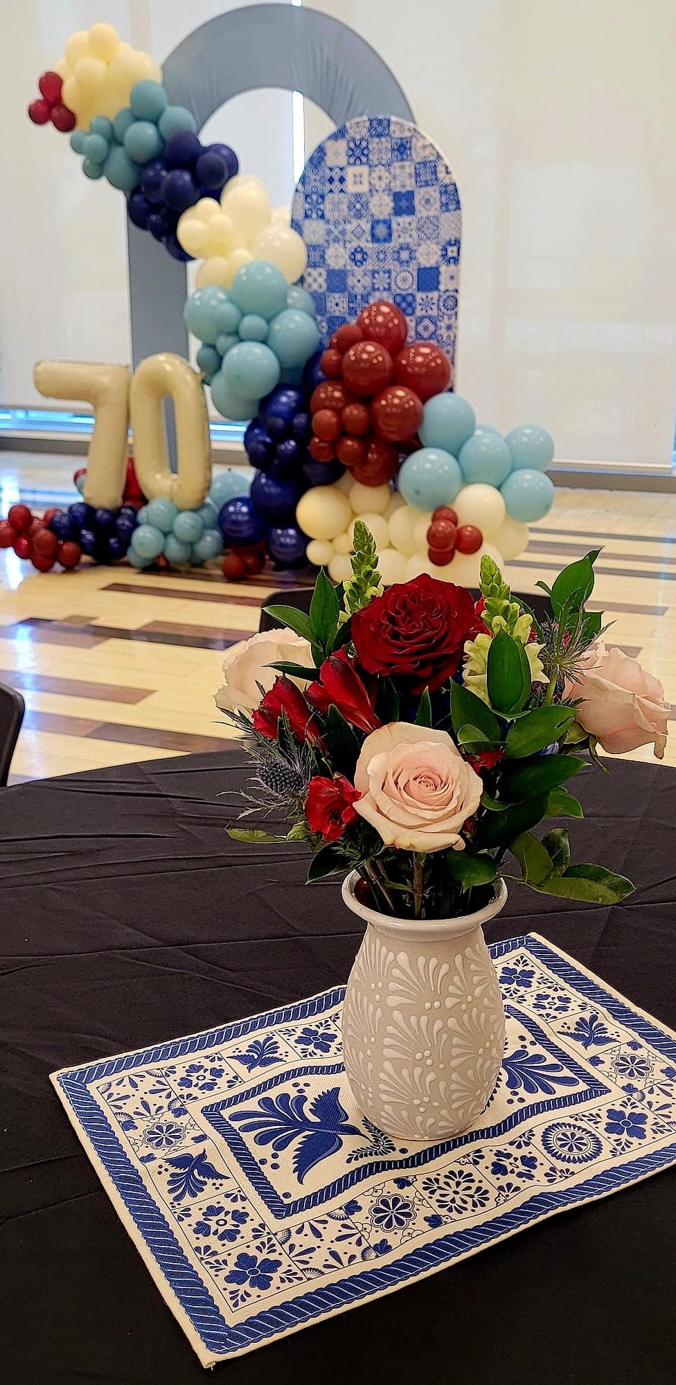 A small arrangement of red and blush roses with greenery in a blue-and-white talavera vase, with a navy, burgundy, light blue, and cream balloon garland forming a 70 backdrop behind it.